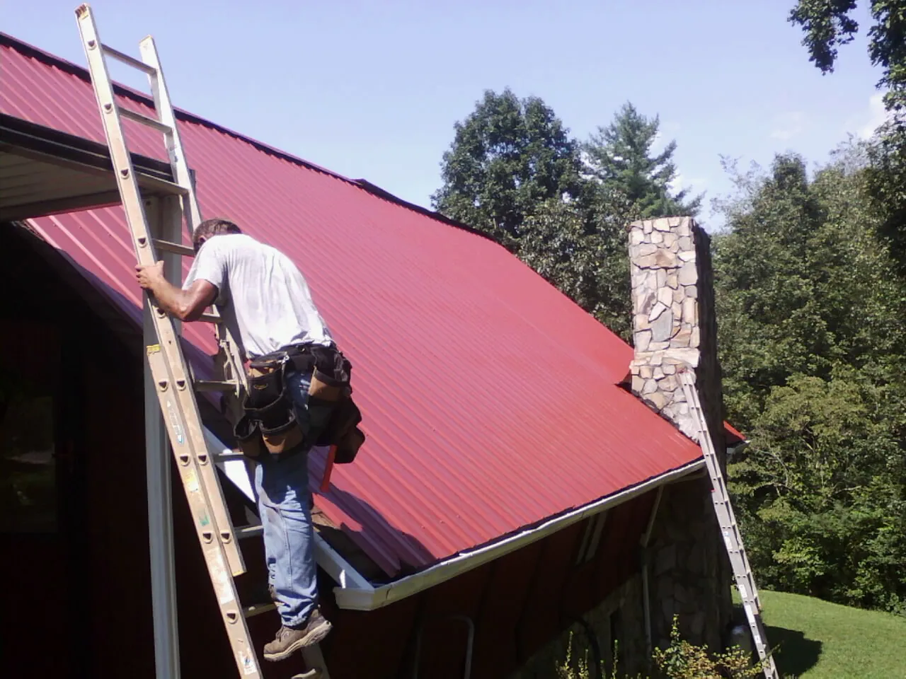 Roofer on a ladder beside a red metal roof and stone chimney — Soffit & Fascia Repair by Roofing Brothers in Tunnelton